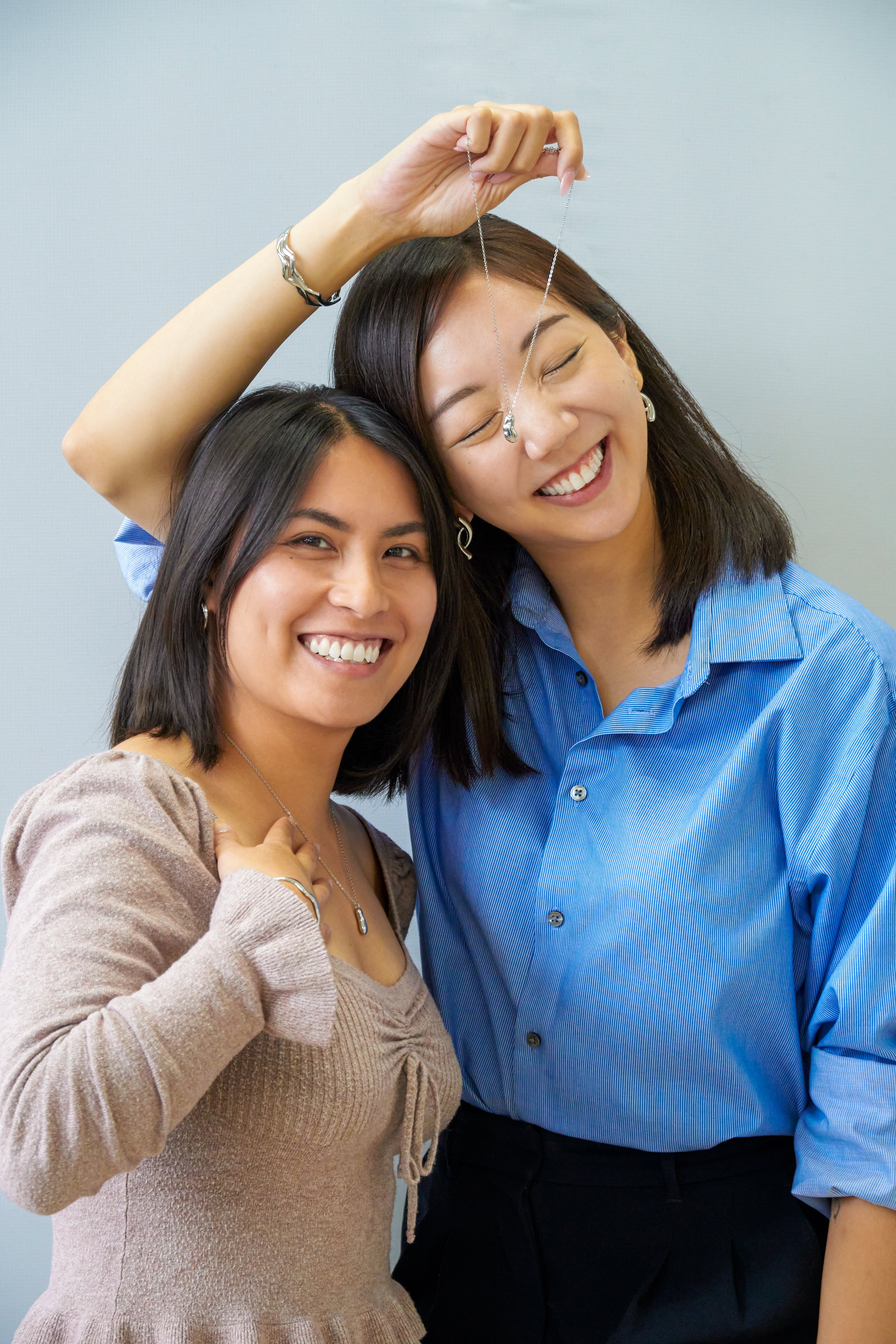 Two women posing together with a plain background and holding jewelry